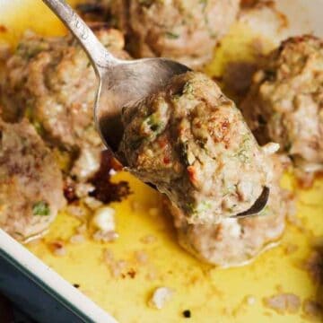 A spoon lifts a baked meatball from a dish filled with several meatballs in a golden, oily sauce.
