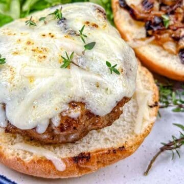 Open-faced cheeseburger on a bun topped with melted cheese and fresh herbs, with caramelized onions and lettuce on the side, served on a white plate with a blue rim.