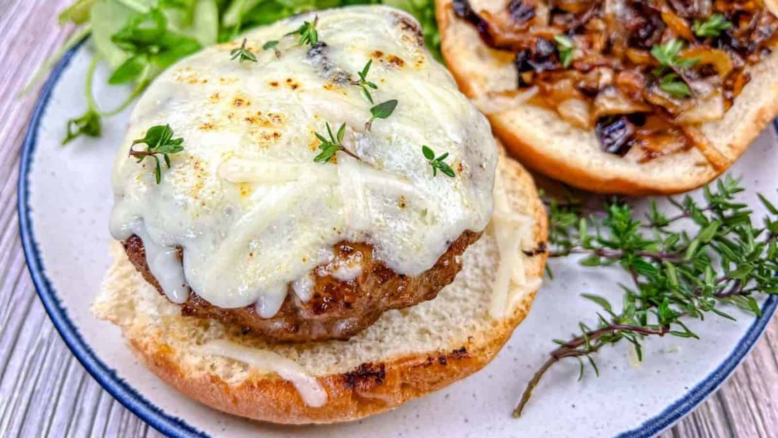 Open-faced cheeseburger on a bun topped with melted cheese and fresh herbs, with caramelized onions and lettuce on the side, served on a white plate with a blue rim.