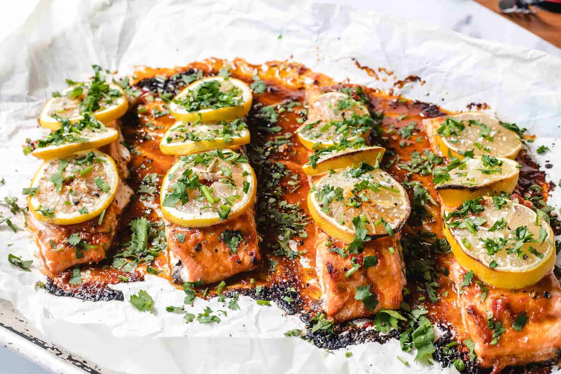 Baked salmon fillets topped with lemon slices and chopped herbs on a parchment-lined baking sheet.