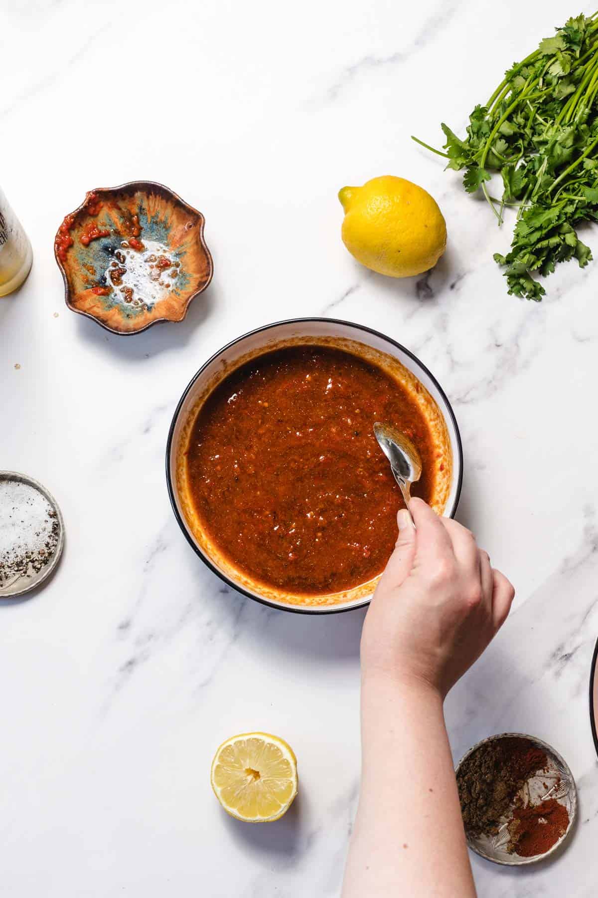 A hand stirs red sauce in a bowl, surrounded by spices, lemon, and fresh herbs on a white surface.