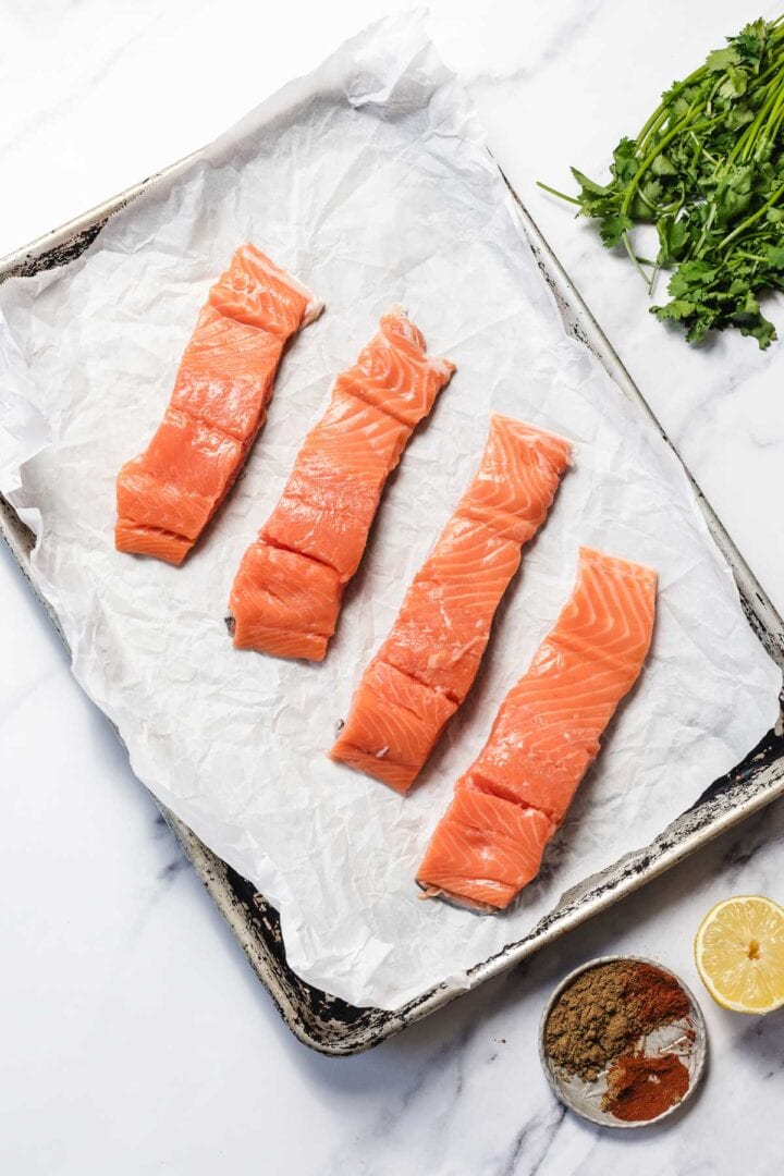 Four raw salmon fillets on a parchment-lined baking sheet with cilantro, lemon, and spices nearby.