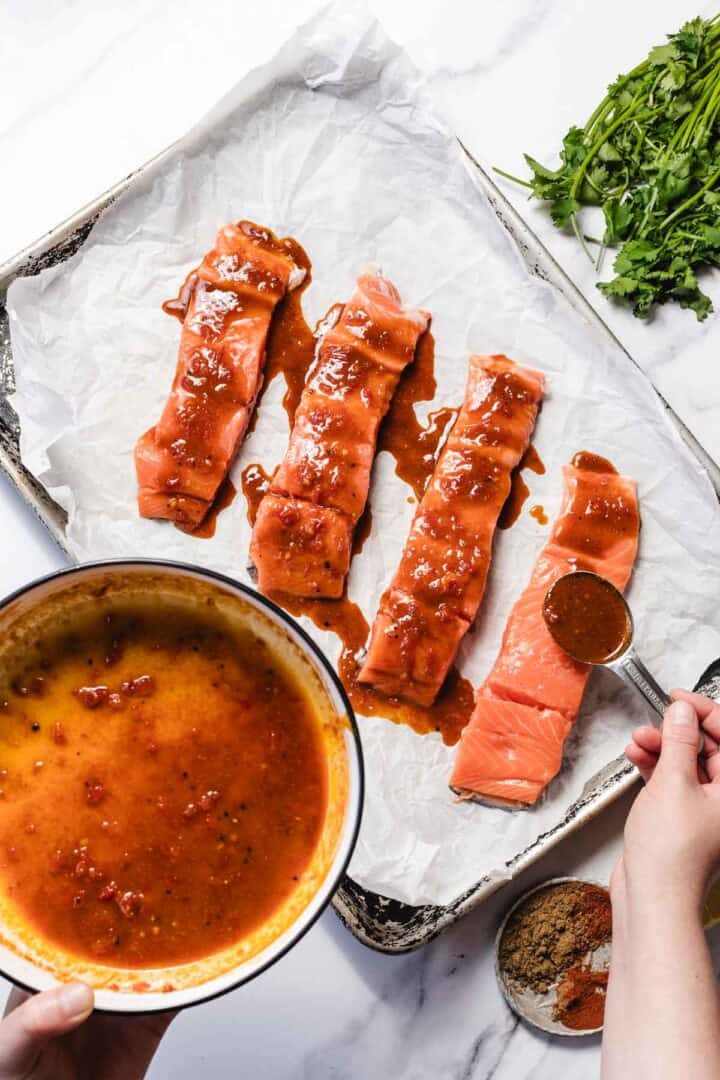 Four raw salmon fillets on parchment, being coated with a reddish marinade from a bowl; cilantro nearby.