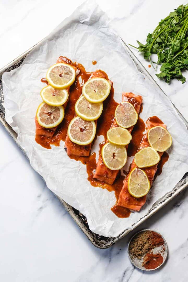 Three salmon fillets topped with lemon slices and sauce on parchment paper, with herbs and spices nearby.