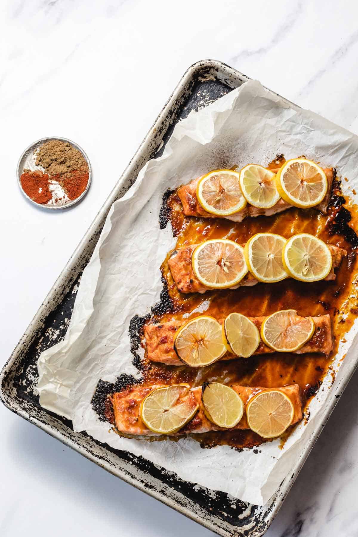 Four baked salmon fillets topped with lemon slices on a parchment-lined baking sheet, with spices in a small bowl.