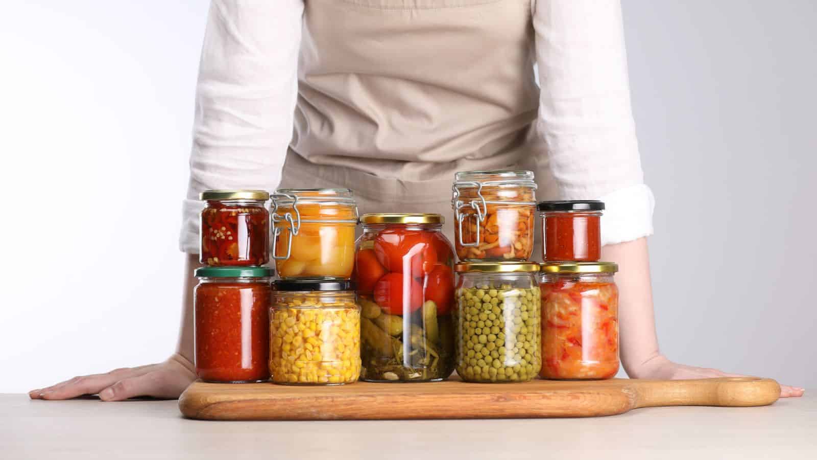 Person in apron stands behind jars of preserved vegetables and fruits on a wooden cutting board, mindful of canning dangers to ensure safe, delicious results.