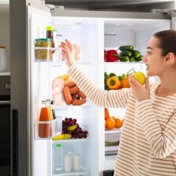 A woman in a striped shirt stands in front of an open refrigerator, holding a green apple and reaching for a jar on the fridge door. The refrigerator is stocked with fruits, vegetables, drinks, and packaged foods.