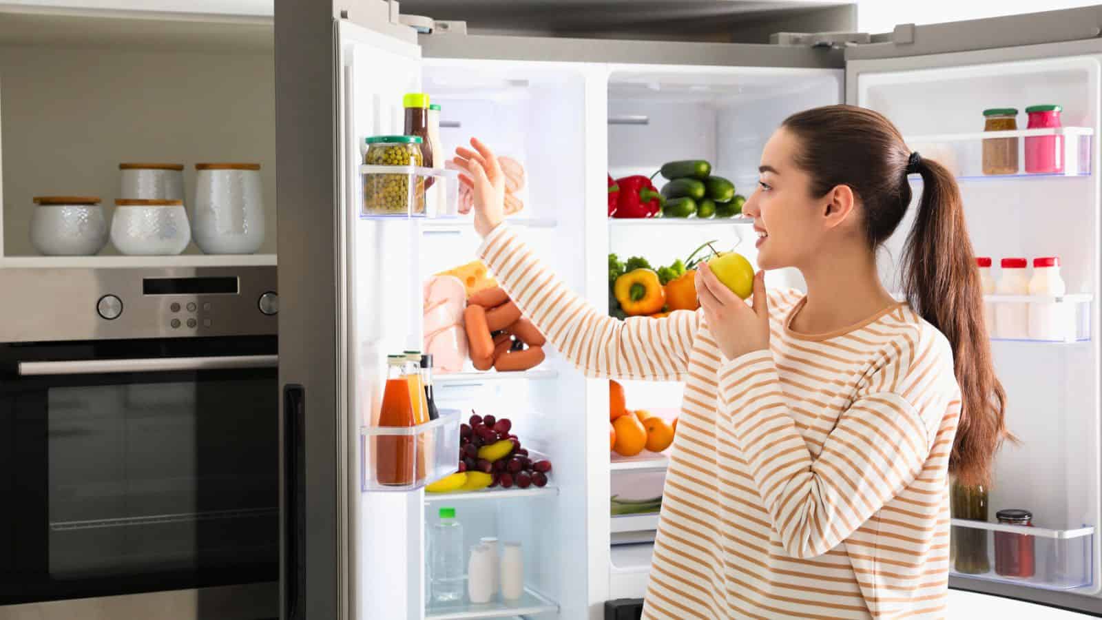 A woman in a striped shirt stands in front of an open refrigerator, holding a green apple and reaching for a jar on the fridge door. The refrigerator is stocked with fruits, vegetables, drinks, and packaged foods.