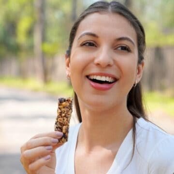 A woman with long brown hair, wearing a white shirt, smiles while holding a granola bar outdoors. The background is blurred with trees and a fence along a pathway.