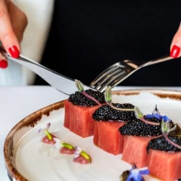 A person with red nail polish uses a fork and knife to cut pieces of watermelon topped with black caviar, garnished with edible flowers, on a white plate with a glass of white wine nearby.