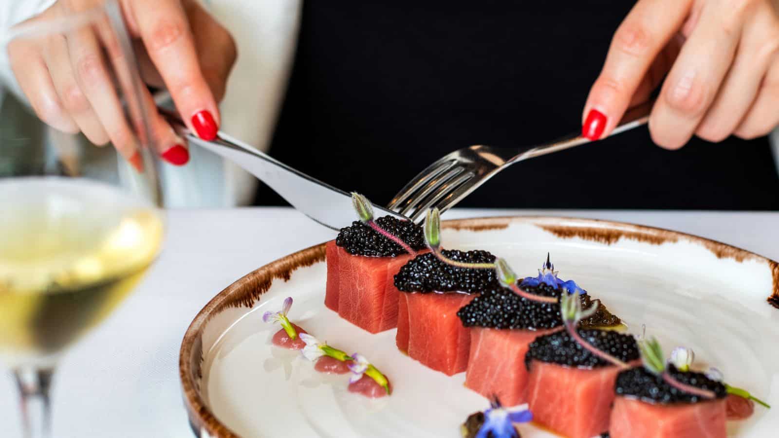 A person with red nail polish uses a fork and knife to cut pieces of watermelon topped with black caviar, garnished with edible flowers, on a white plate with a glass of white wine nearby.