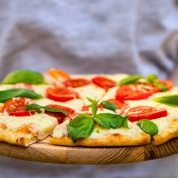 A person holding a wooden board with a pizza topped with melted cheese, tomato slices, and fresh basil leaves. The person is wearing a light gray shirt and the background is blurred.