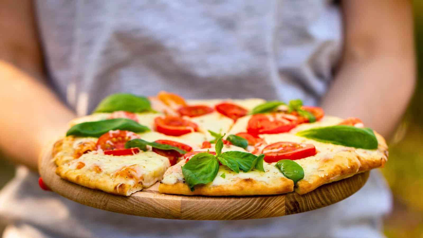 A person holding a wooden board with a pizza topped with melted cheese, tomato slices, and fresh basil leaves. The person is wearing a light gray shirt and the background is blurred.