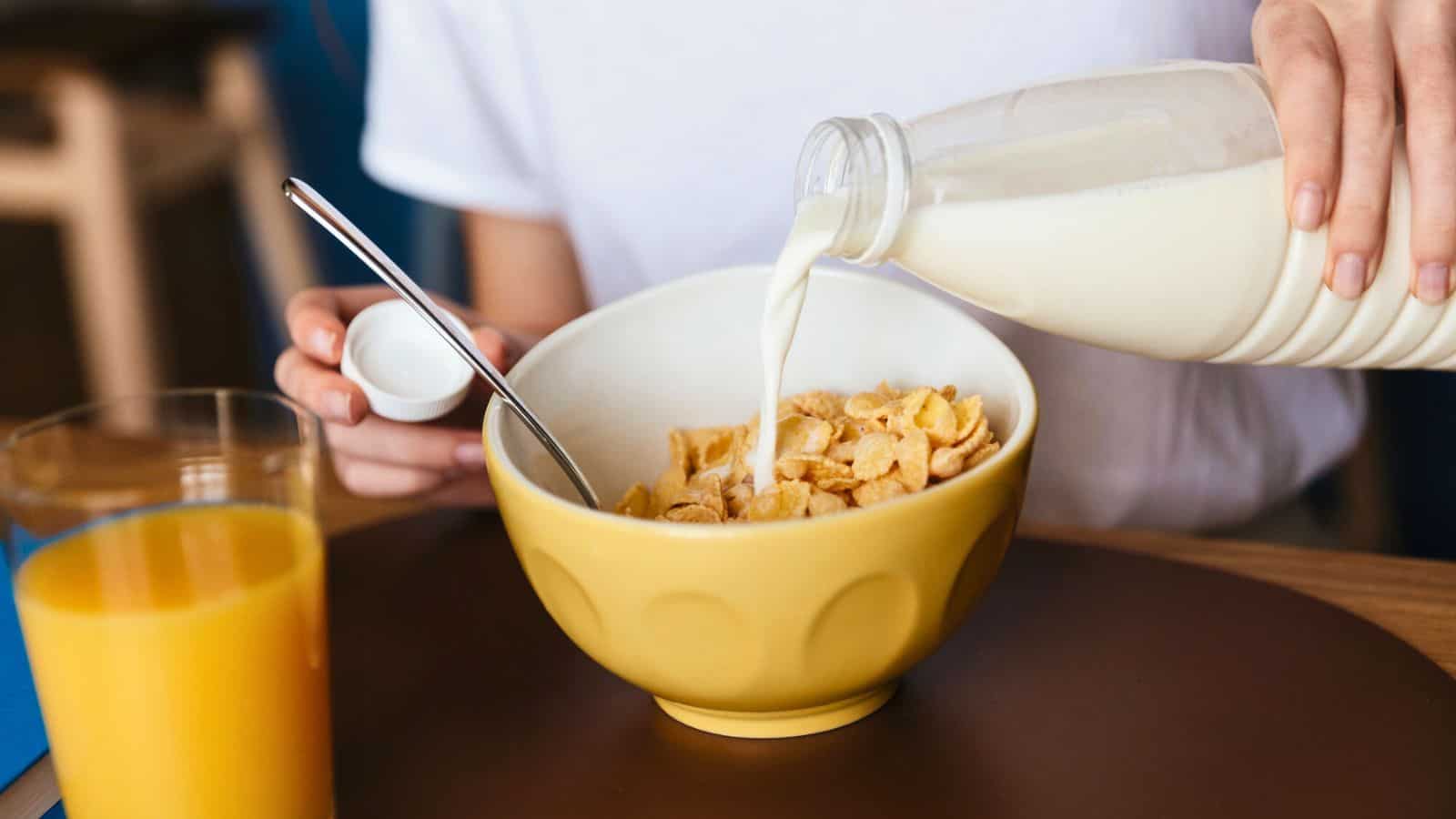A person pours milk from a bottle into a yellow bowl filled with cornflakes. A spoon rests in the bowl, and a glass of orange juice sits on the table next to it.