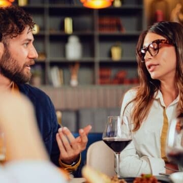 A man and a woman sit at a restaurant table, talking and drinking wine.