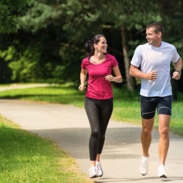 A man and woman jogging together on a paved path in a green, sunny park, smiling at each other.