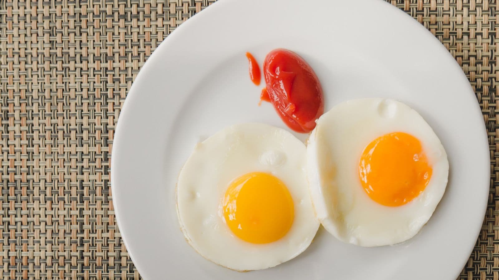 Two sunny-side-up eggs and a serving of ketchup on a white plate, placed on a woven placemat.