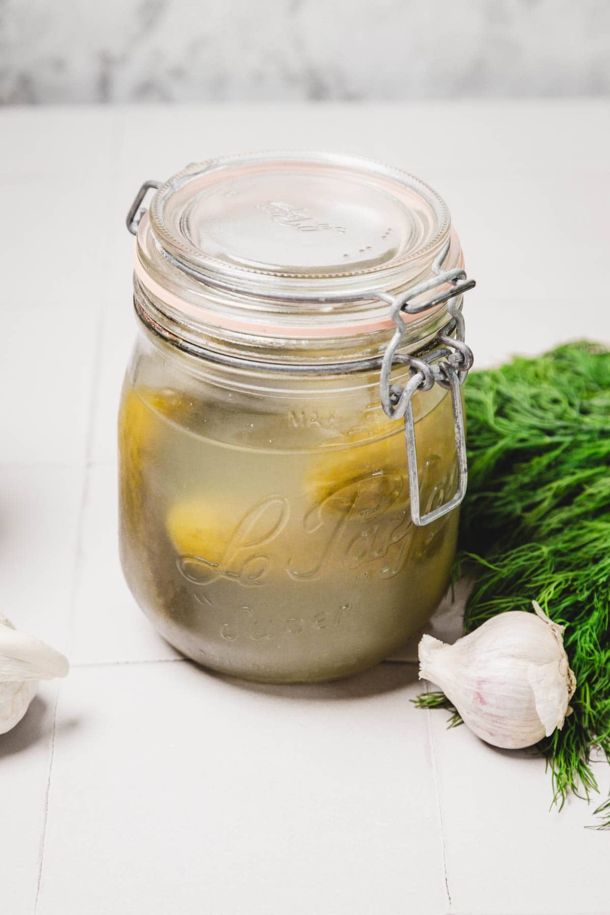 A glass jar of pickles beside fresh garlic and dill on a white surface.
