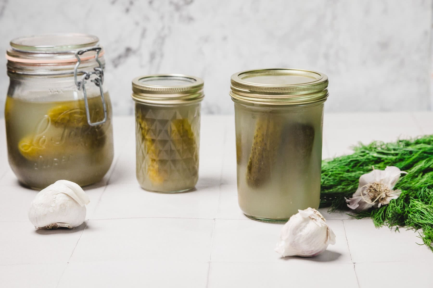 Three jars of pickles with garlic cloves and fresh dill on a white surface.