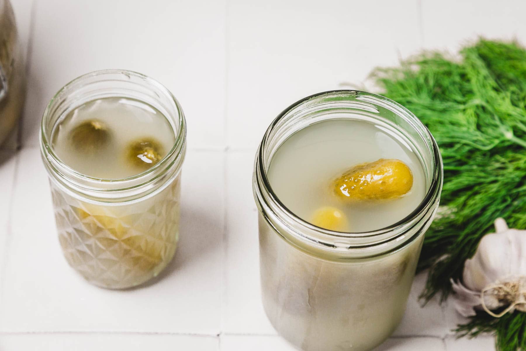 Two jars of pickles in brine on a white surface, with fresh dill and garlic nearby.
