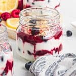 A glass jar filled with layered yogurt, lemon berry coconut custard, and granola on a light surface, with a napkin nearby.