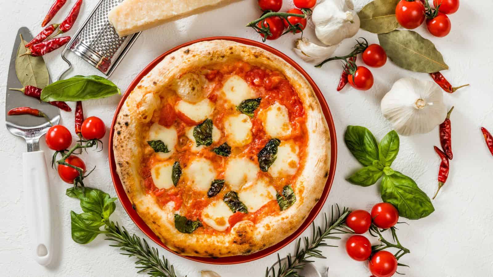 A Margherita pizza with melted mozzarella, tomato sauce, and basil sits on a plate, surrounded by cherry tomatoes, garlic, red chili peppers, fresh basil, rosemary, and a block of cheese with a grater.
