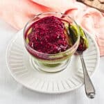 Glass of green pudding topped with raspberry sauce, served with a spoon on a white plate.