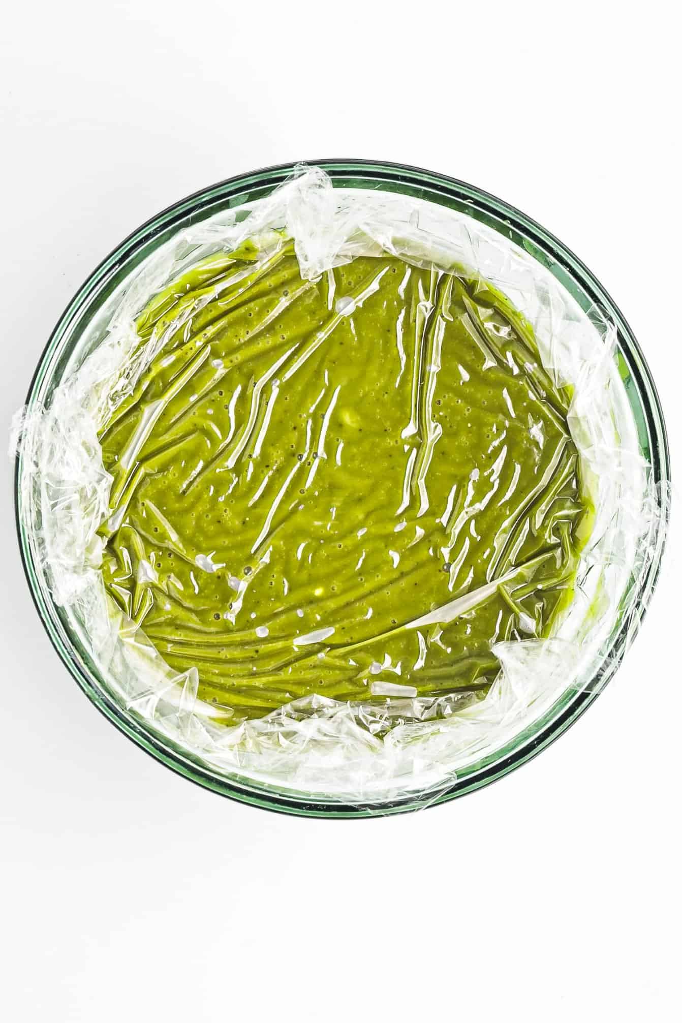 A bowl of green batter covered with plastic wrap, viewed from above on a white background.