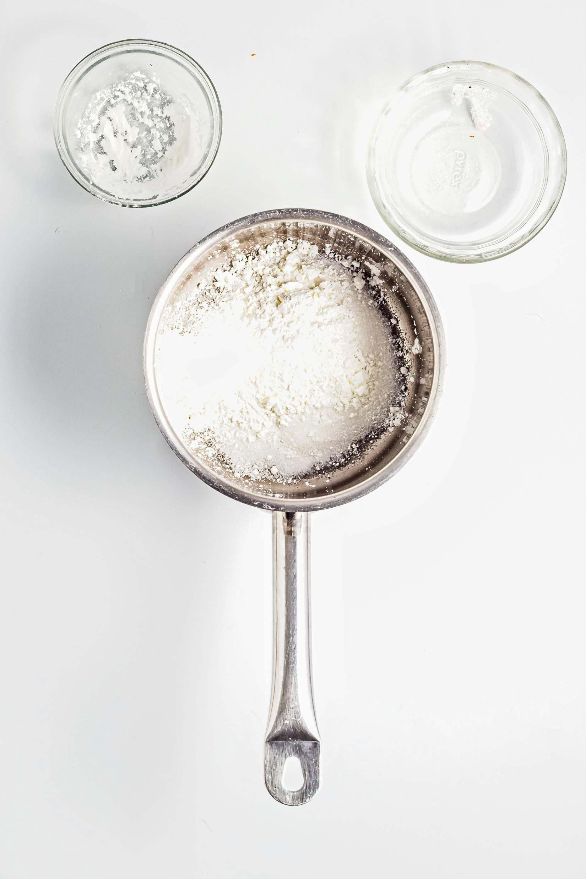 A saucepan with white powder, next to two small glass bowls on a white surface.