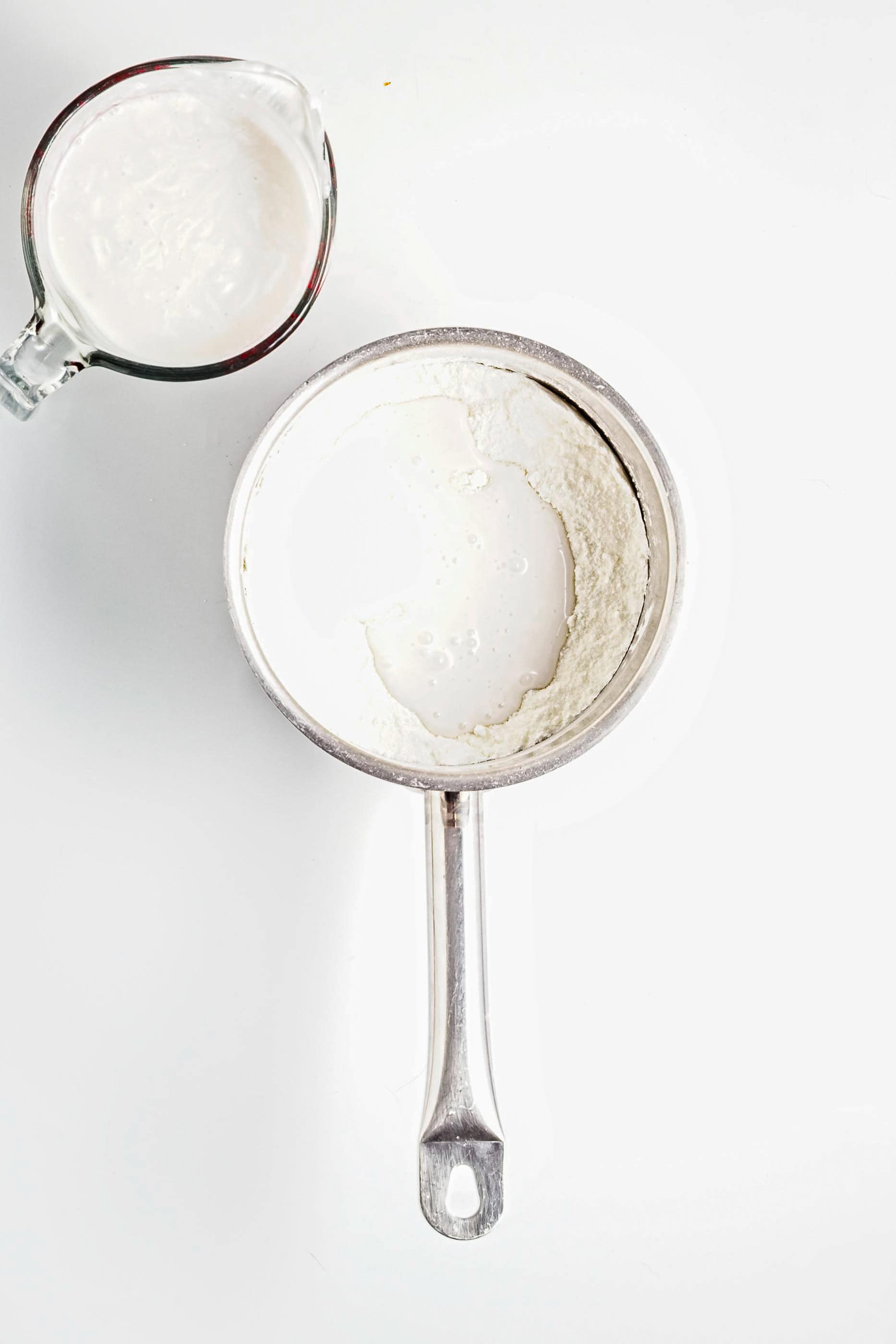 A saucepan with a creamy mixture and a measuring cup filled with milk on a white surface.