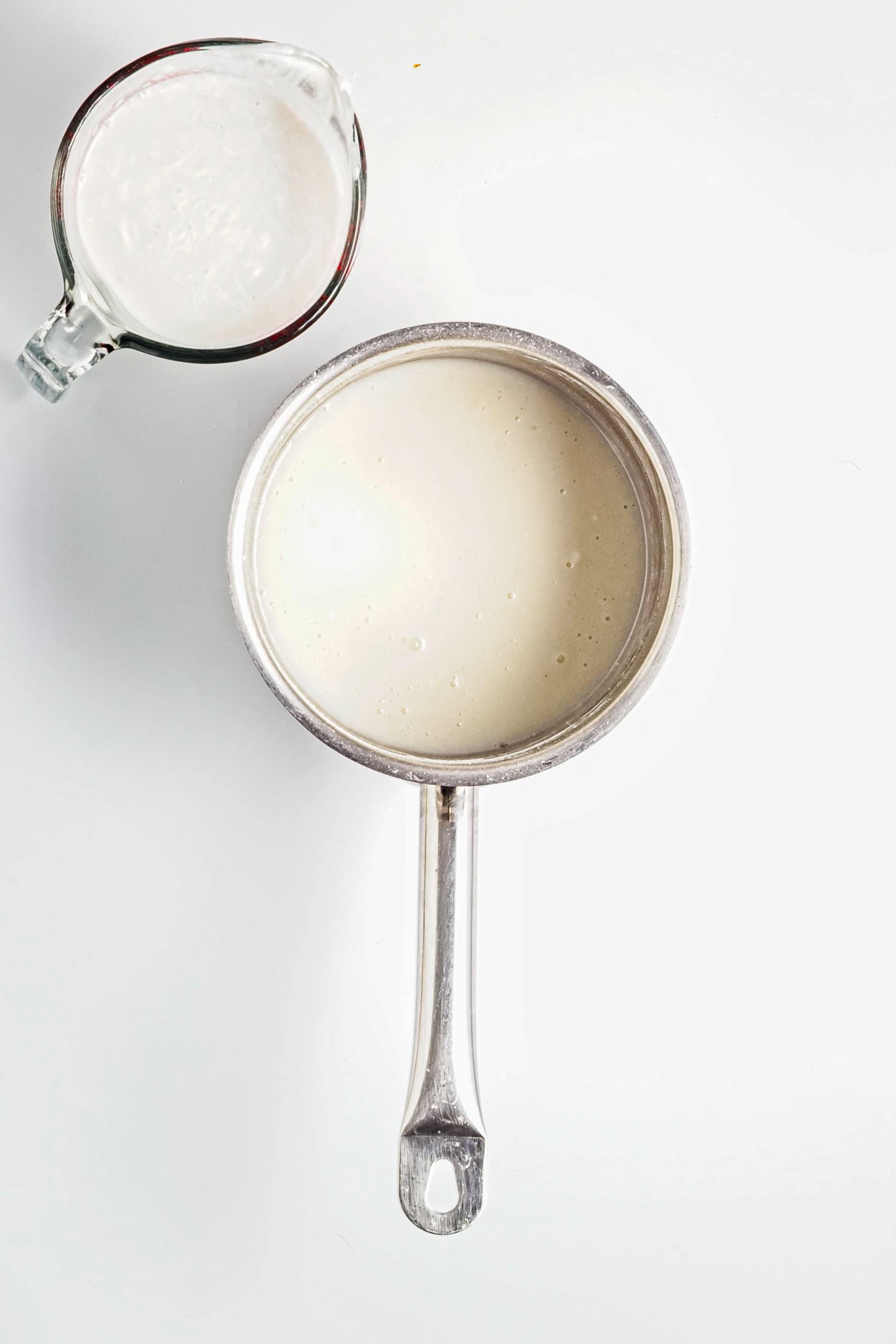 A metal saucepan with white liquid next to a glass measuring cup with a similar white mixture.