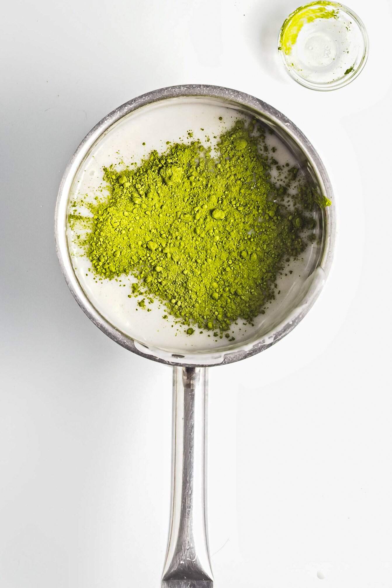Overhead view of a saucepan with milk and matcha powder, next to a small glass dish with matcha residue.