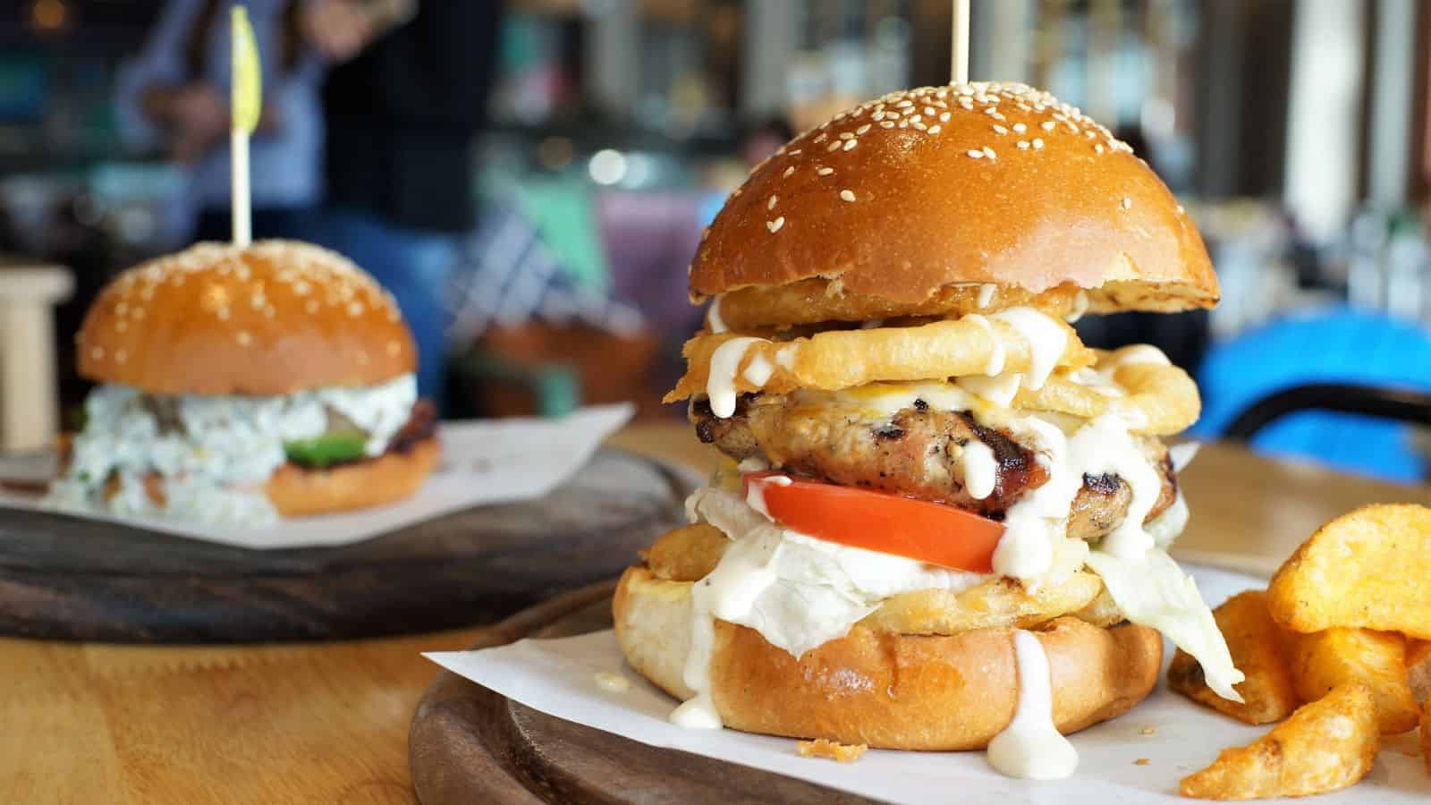 A close-up of a chicken burger with lettuce, tomato, onion rings, and creamy sauce in a sesame bun, served on a wooden board with potato chips. Another burger is blurred in the background.