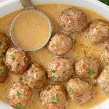 Meatballs in creamy brown sauce, garnished with parsley, served in a white oval dish with a ladle.