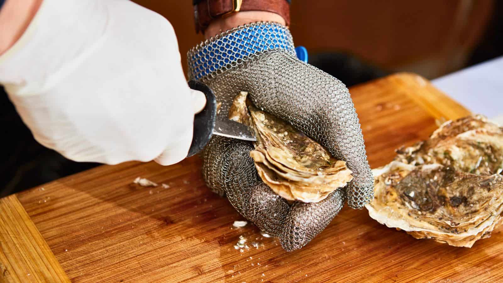 A person wearing a metal mesh glove holds an oyster on a wooden board while using a shucking knife to open it. Another hand, covered with a white glove, steadies the oyster.