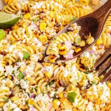 A bowl of creamy pasta salad with corn, cheese, red onion, herbs, and lime wedges, served with wooden utensils.