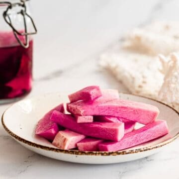 A plate of pink pickled vegetable sticks next to a jar of pink liquid on a marble surface.