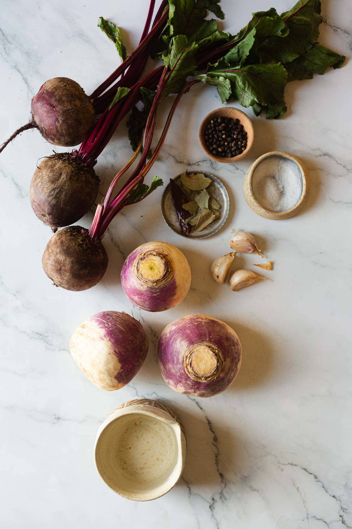 Three beets, three turnips, garlic, bay leaves, peppercorns, salt, and a small ceramic pot on a marble surface—classic ingredients for Middle Eastern Pickled Turnips.