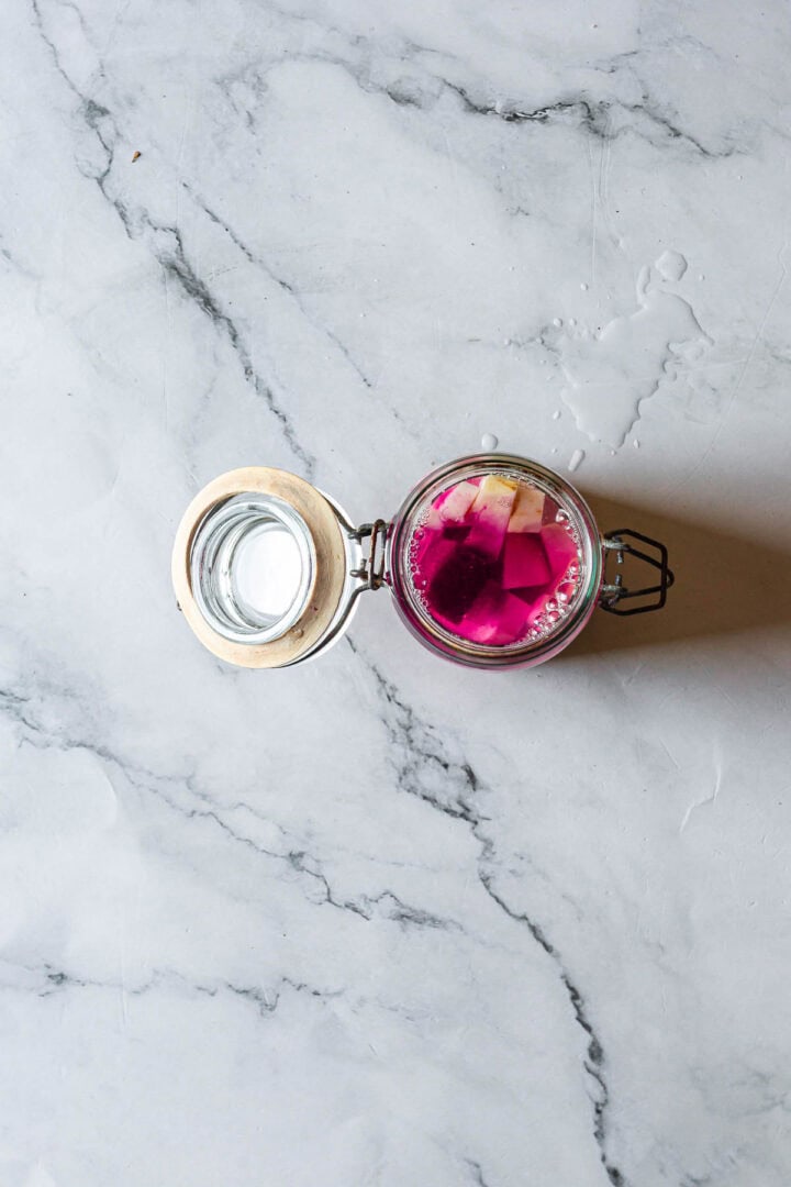 Open glass jar of Middle Eastern Pickled Turnips with pink liquid and white cubes on a marble surface, viewed from above.