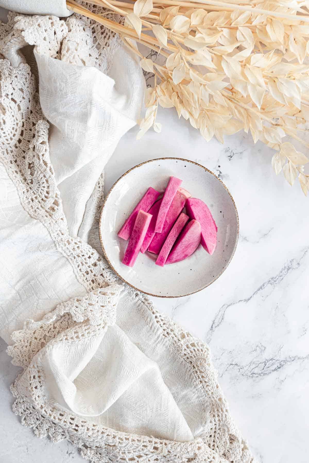 A plate of sliced purple sweet potato on a white cloth with dried beige leaves beside it, reminiscent of the vibrant hues found in Middle Eastern Pickled Turnips.