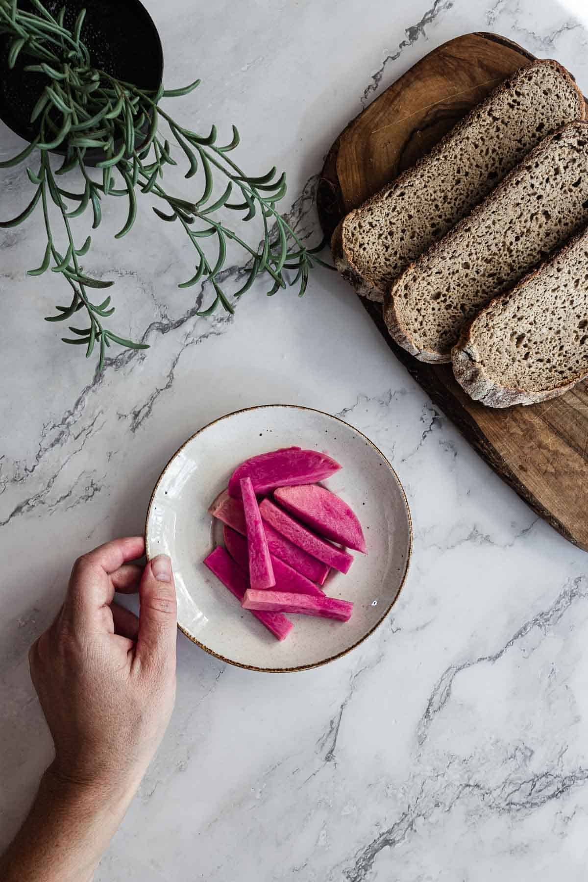 Hand holding plate of Middle Eastern Pickled Turnips, with rustic bread and rosemary on marble surface.