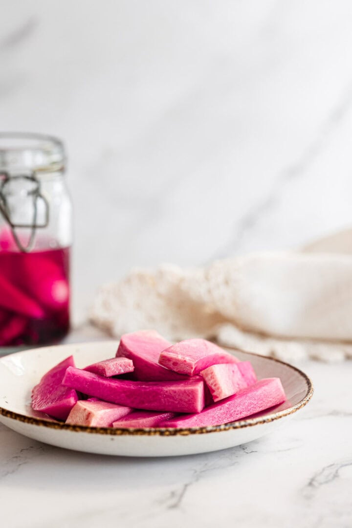 Sliced pink Middle Eastern Pickled Turnips on a plate, with a jar of more pickled turnips in the background.