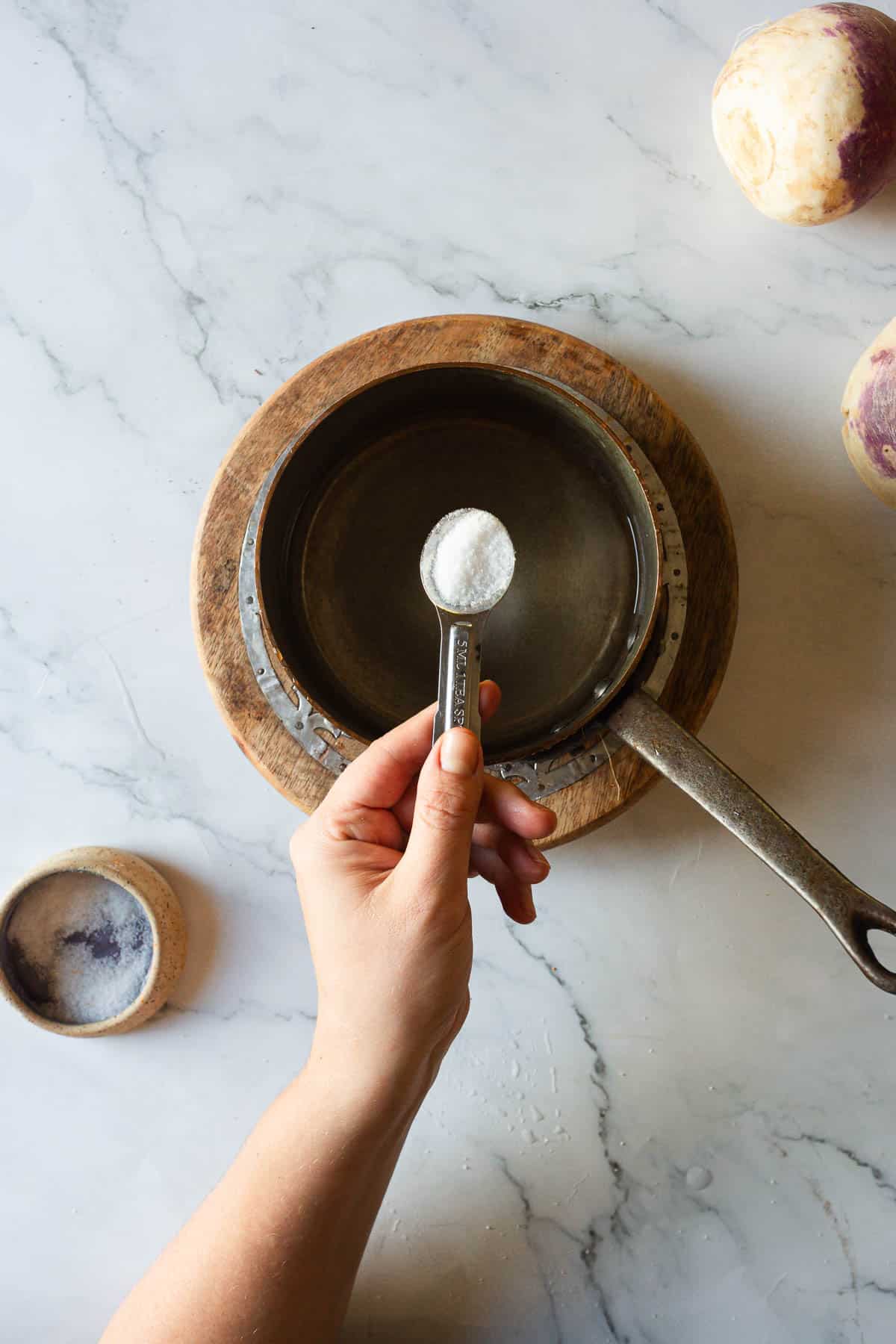 A hand holds a spoon of salt above an empty saucepan on a wooden board, ready to prepare Middle Eastern Pickled Turnips, with a bowl of salt nearby.