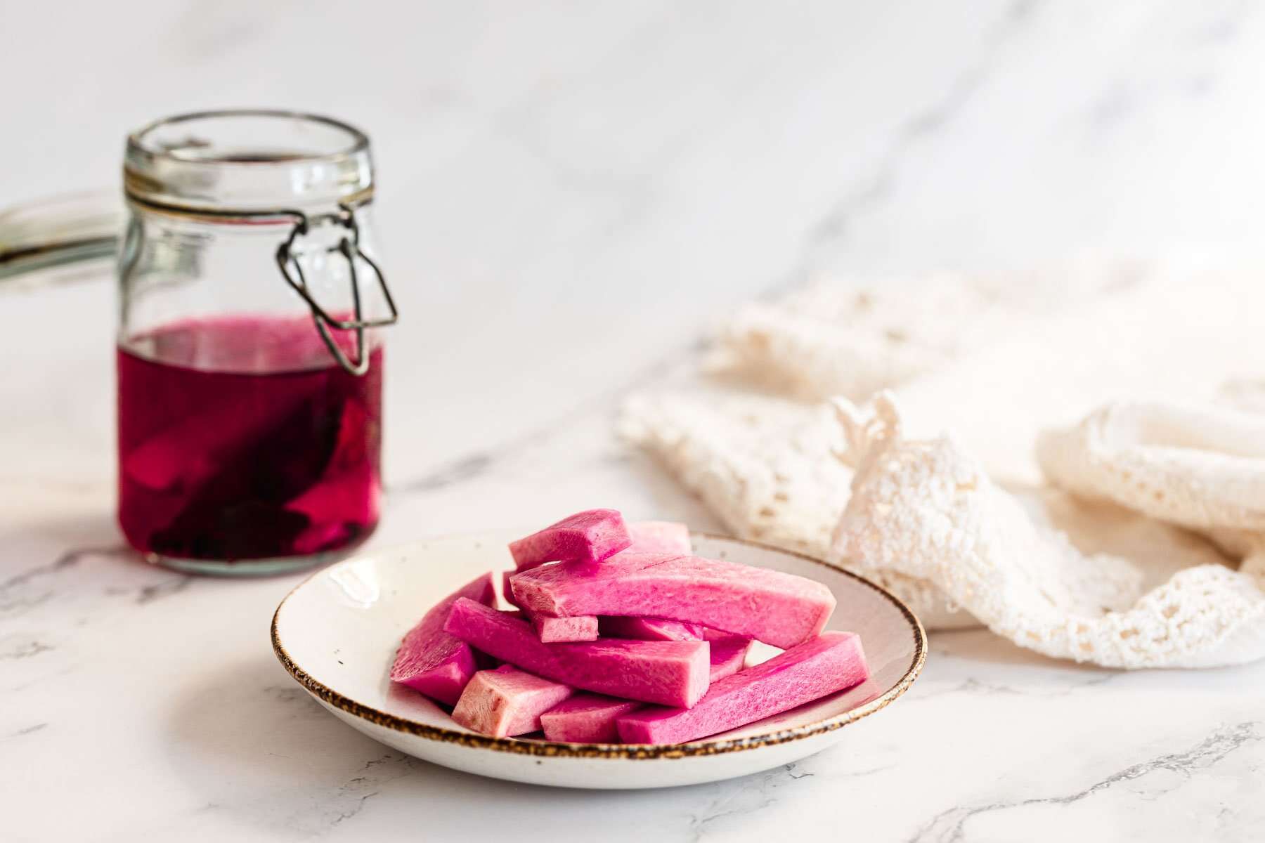 Jar of pink liquid and a plate of Middle Eastern Pickled Turnips on a white marble surface.