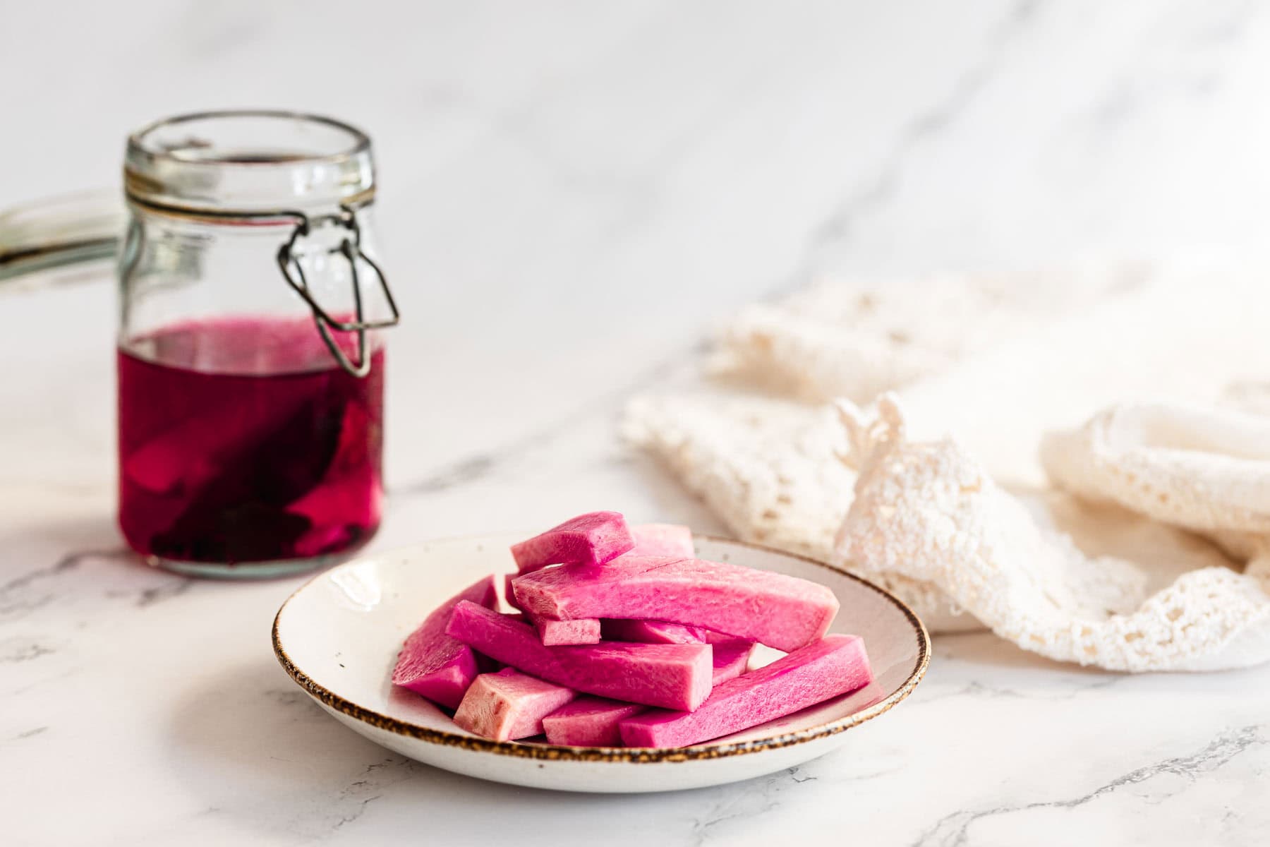 Plate of pink pickled vegetables, including Middle Eastern Pickled Turnips, next to a jar of vibrant pink brine on a white marble surface.