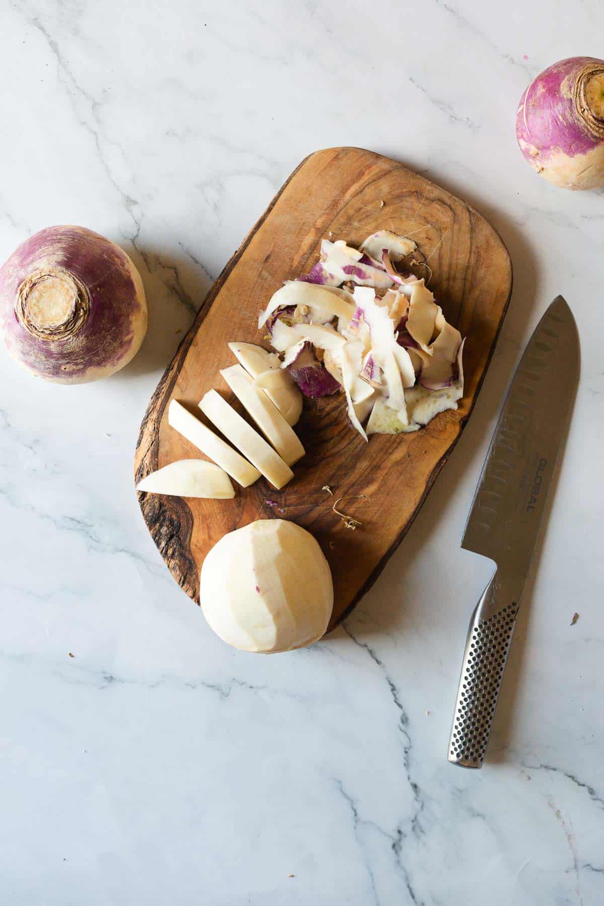 A peeled and sliced turnip on a wooden board with peelings, a knife, and whole turnips on a marble surface, ready for making Middle Eastern Pickled Turnips.