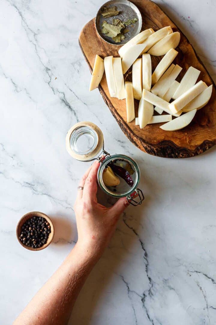 A hand holds an open jar with spices beside sliced parsnips, bay leaves, and peppercorns on a marble surface, evoking flavors reminiscent of Middle Eastern Pickled Turnips.