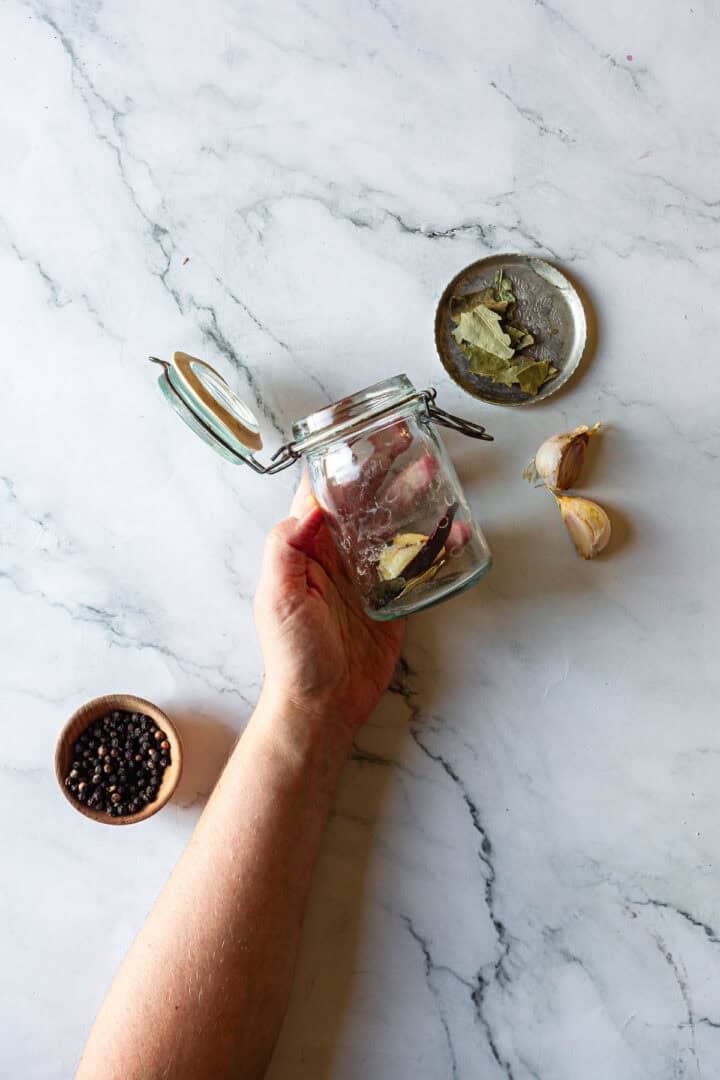 A hand holds an empty glass jar with bay leaves; peppercorns, garlic, and more bay leaves are nearby on a marble surface—perfect for starting Middle Eastern Pickled Turnips.