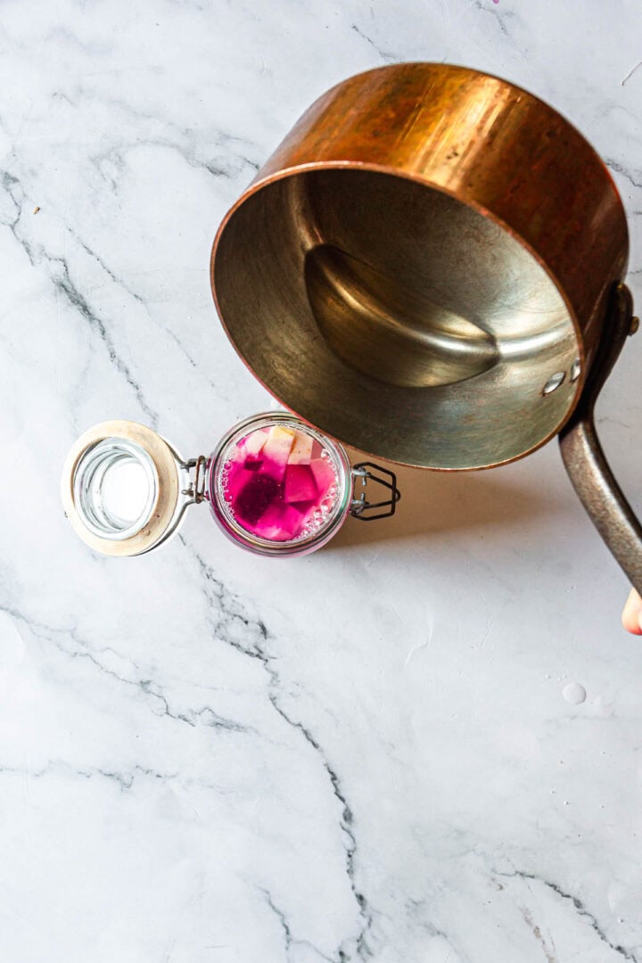 A saucepan is being tilted above a glass jar filled with pink liquid and radish slices, resembling the vibrant brine used for Middle Eastern Pickled Turnips, on a marble surface.