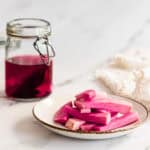 A plate of bright pink Middle Eastern Pickled Turnips sits next to a jar of vibrant pink pickling liquid on a white surface.